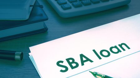 A piece of paper that says "SBA Loan" lies on a desk. There is a calculator, two notebooks, and a pen near the paper on the desk.