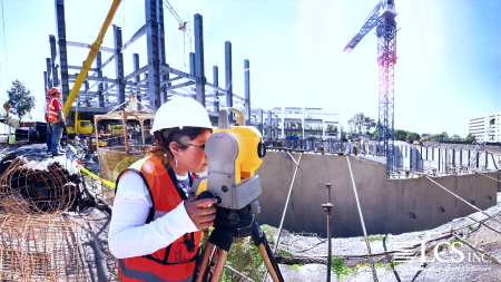 A female construction worker in safety gear uses a theodolite to survey a building site, with steel framework and cranes visible in the background.
