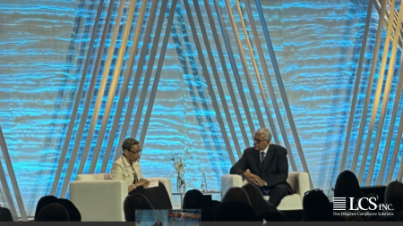 Two speakers in a fireside chat on a conference stage with a blue geometric backdrop, audience silhouettes in the foreground.