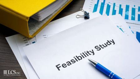 A printed feasibility study document on a desk alongside financial charts, a blue pen, and a yellow binder, representing project analysis and due diligence.