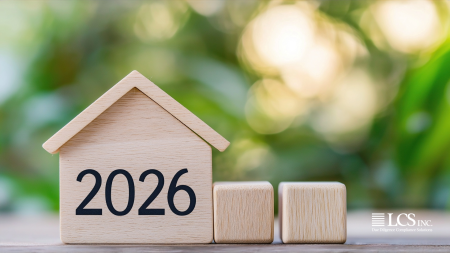 Wooden house‑shaped block displaying ‘2026’ beside two blank wooden cubes, with a soft green blurred background and the LCS Inc. logo in the corner.