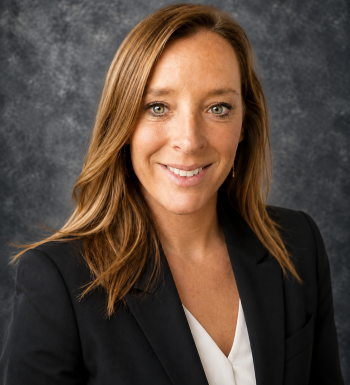 Professional headshot of Liz Mahoney smiling in a dark blazer and white blouse against a textured gray studio background.